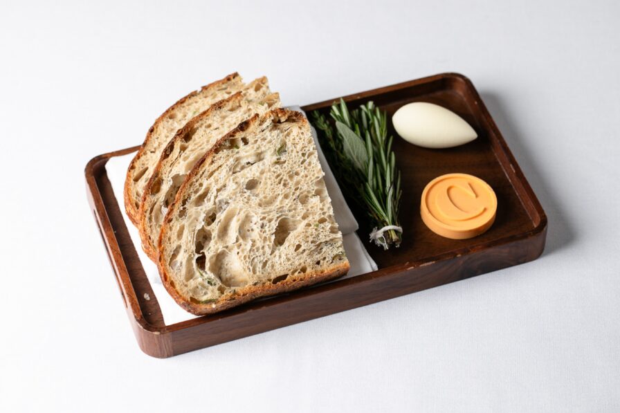 On a white tablecloth, a wooden tray features three slices of bread, a sprig of rosemary, a white oval object, and a round orange disc—an elegant representation of Atlanta's upscale dining scene.