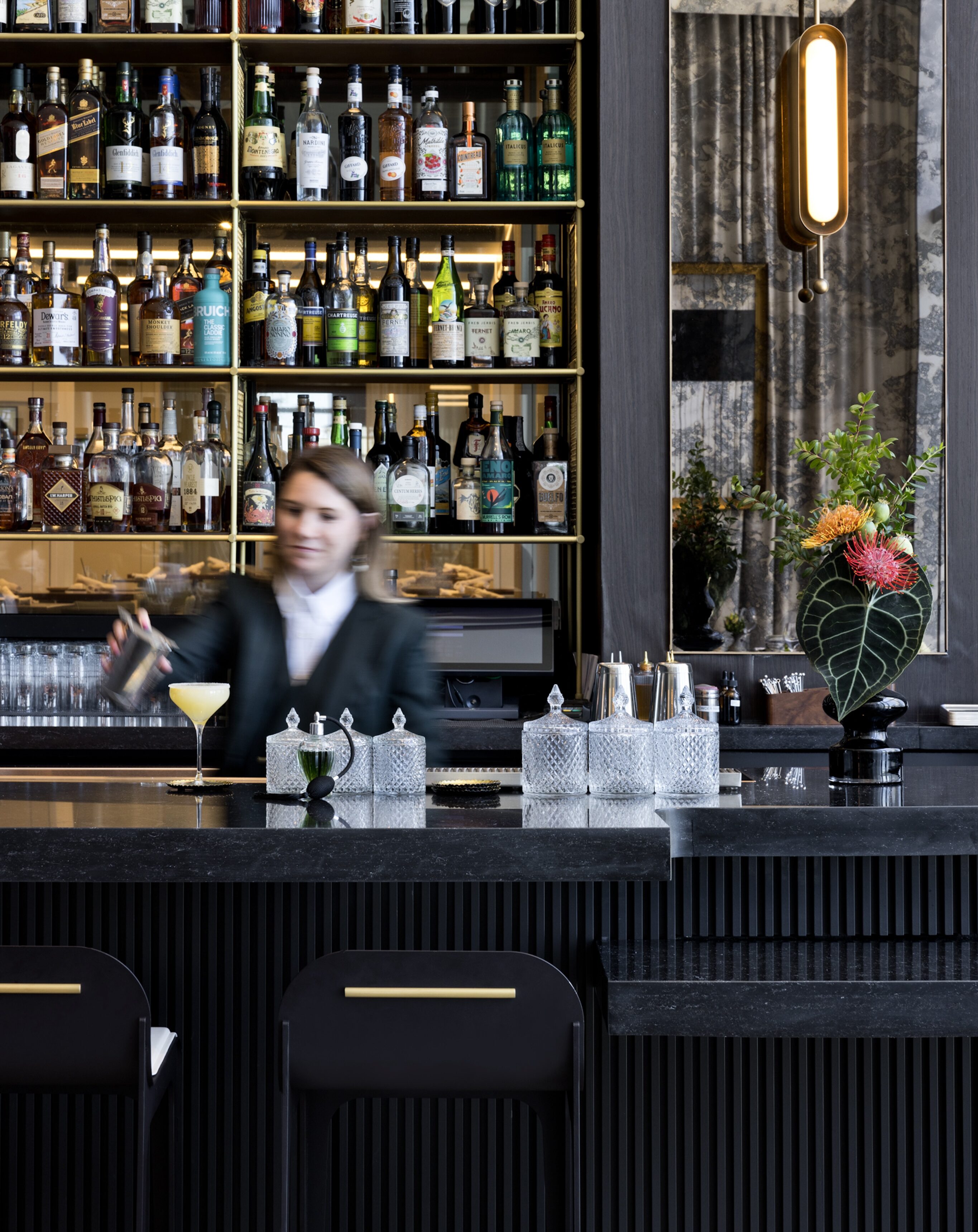 An upscale bartender expertly mixes a drink behind a polished counter with a yellow cocktail glass, surrounded by several bottles on shelves. The Atlanta bar boasts decorative elements reminiscent of Italian dining and elegant lighting to create an inviting atmosphere.