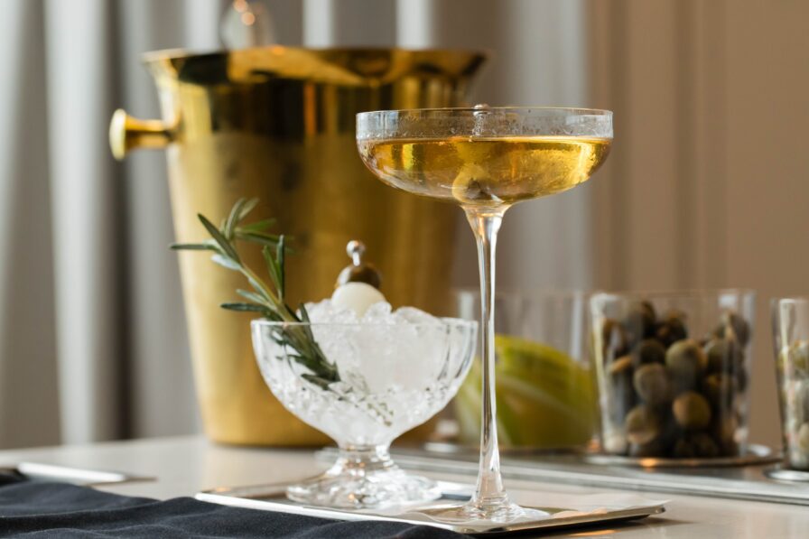 A champagne coupe with a drink and olive garnish is placed next to a crystal bowl of ice with rosemary, capturing the essence of an upscale Atlanta restaurant. A gold ice bucket glimmers softly in the background.