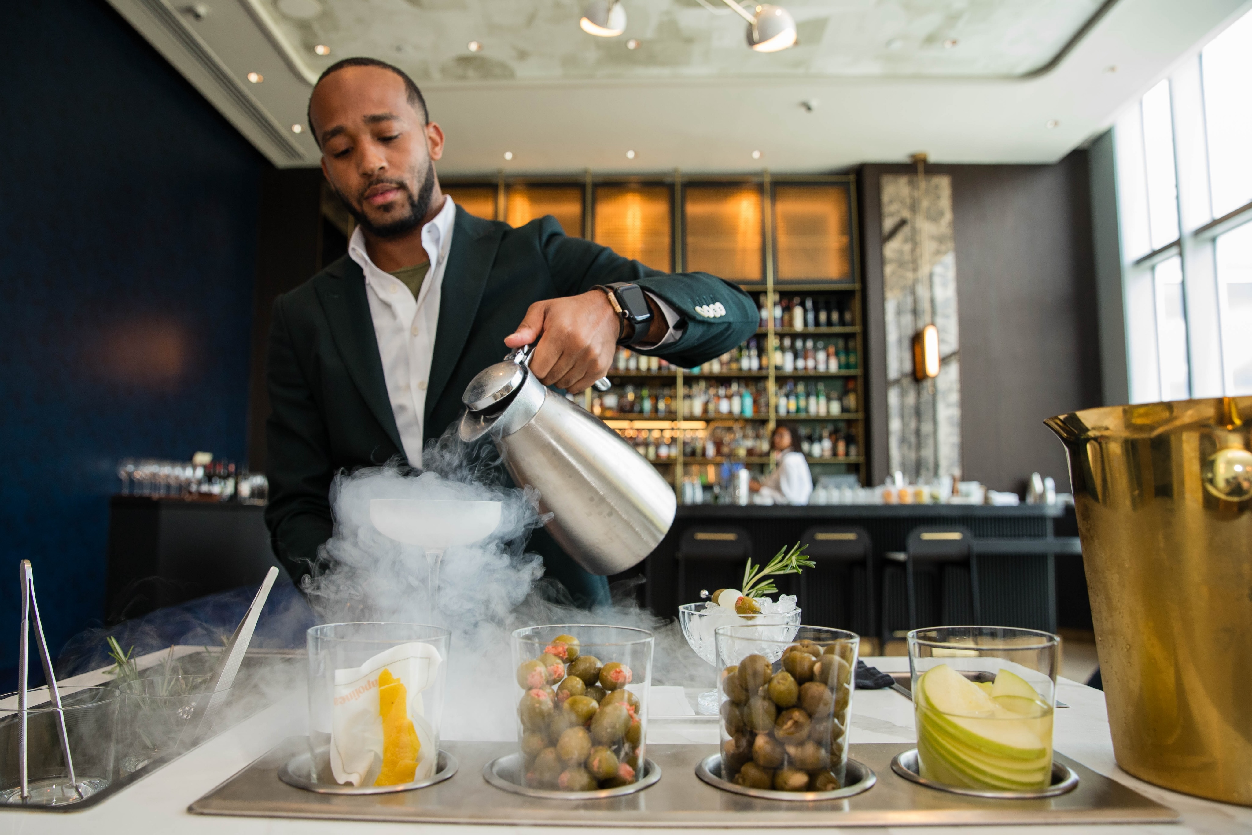 At an upscale bar counter, a person pours liquid nitrogen while olives, citrus slices, and grapes are elegantly displayed in containers, capturing the essence of fine Italian dining.