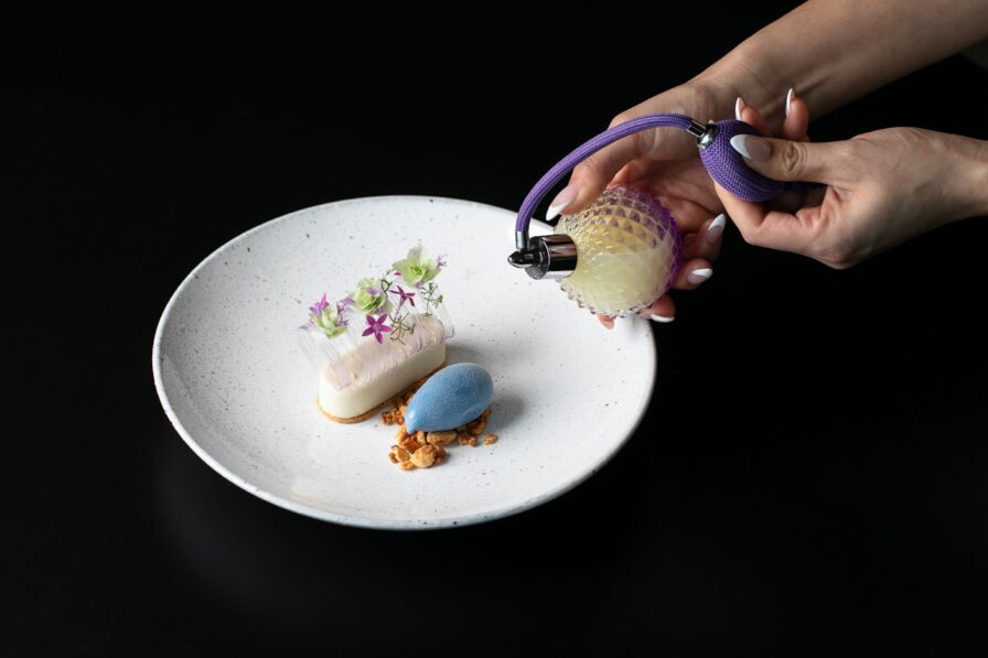 A person delicately sprays a glass of liquid onto a plated dessert, showcasing a velvety white mousse, a striking blue sphere, delicate flowers, and fine crumbs on a pristine plate. This elegant creation epitomizes upscale Italian dining in the heart of Atlanta against an exquisite black backdrop.