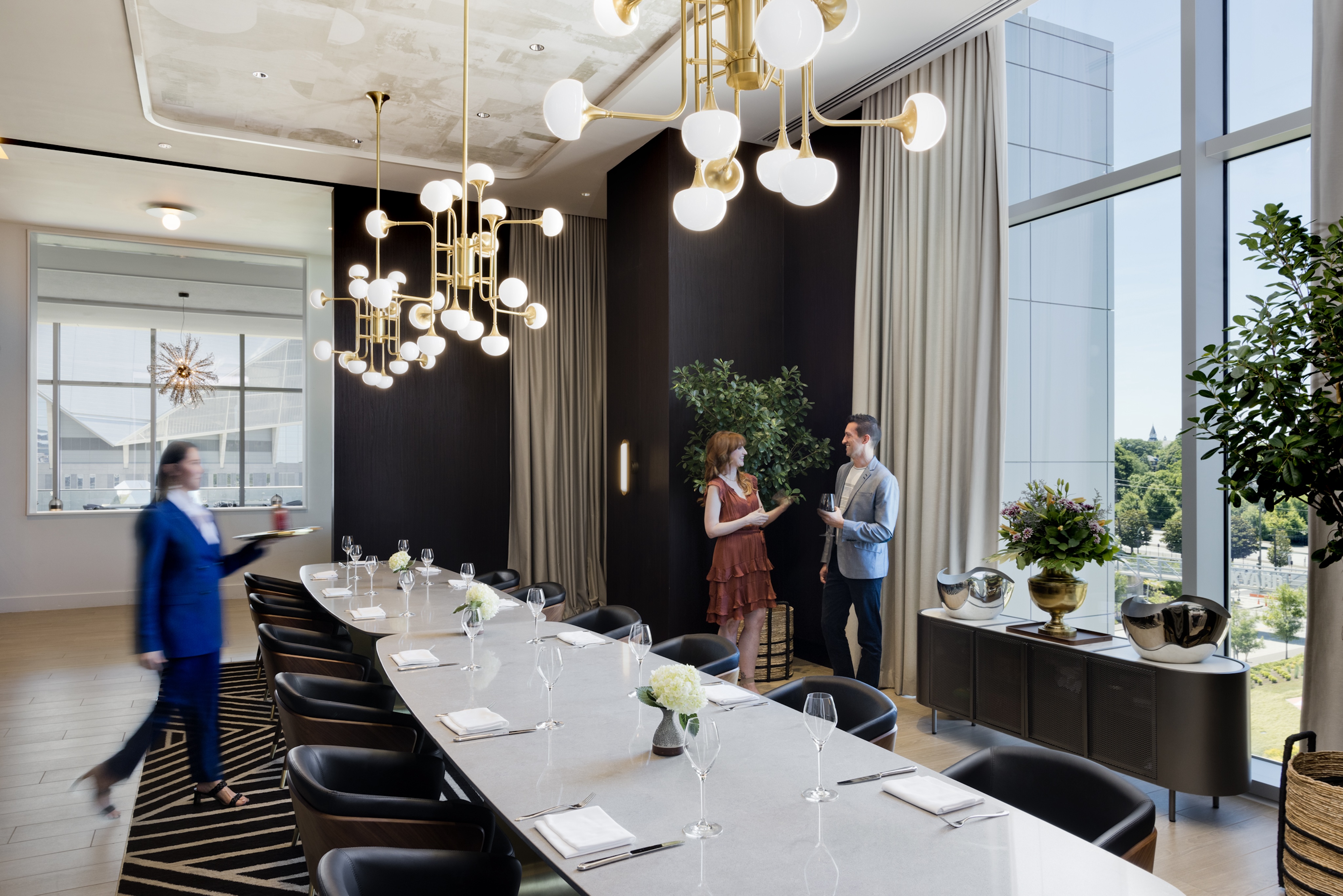 In an upscale dining room, a modern setting features an elegantly set long table. Two people converse while a server from the Italian restaurant walks by, and large windows reveal a lush view of greenery outside.