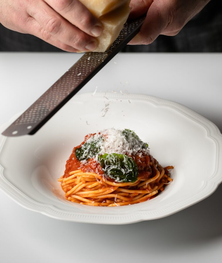 In an ambiance of Italian dining, a person delicately grates cheese over a plate of spaghetti adorned with tomato sauce and greens, all elegantly presented on a white plate.