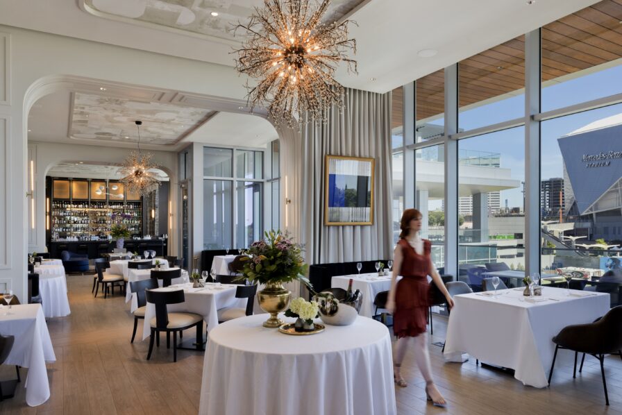 A woman in a red dress strolls through an upscale Atlanta restaurant, past tables set with white tablecloths, illuminated by natural light streaming through large windows and the glow of an ornate chandelier.