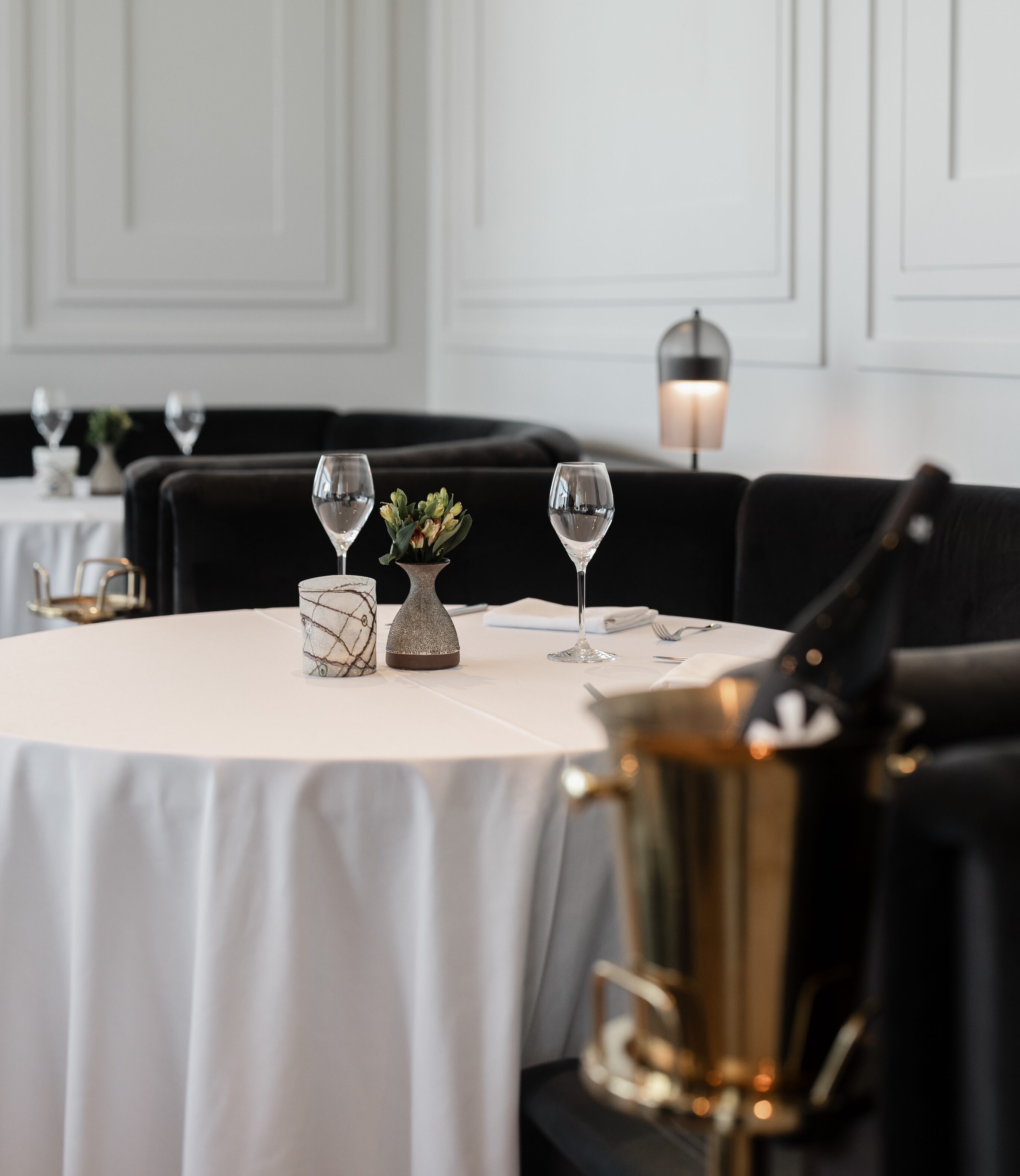 Upscale dining setup with round tables, white tablecloths, wine glasses, small potted plants, and a champagne bucket in a softly lit room reminiscent of fine Italian dining.