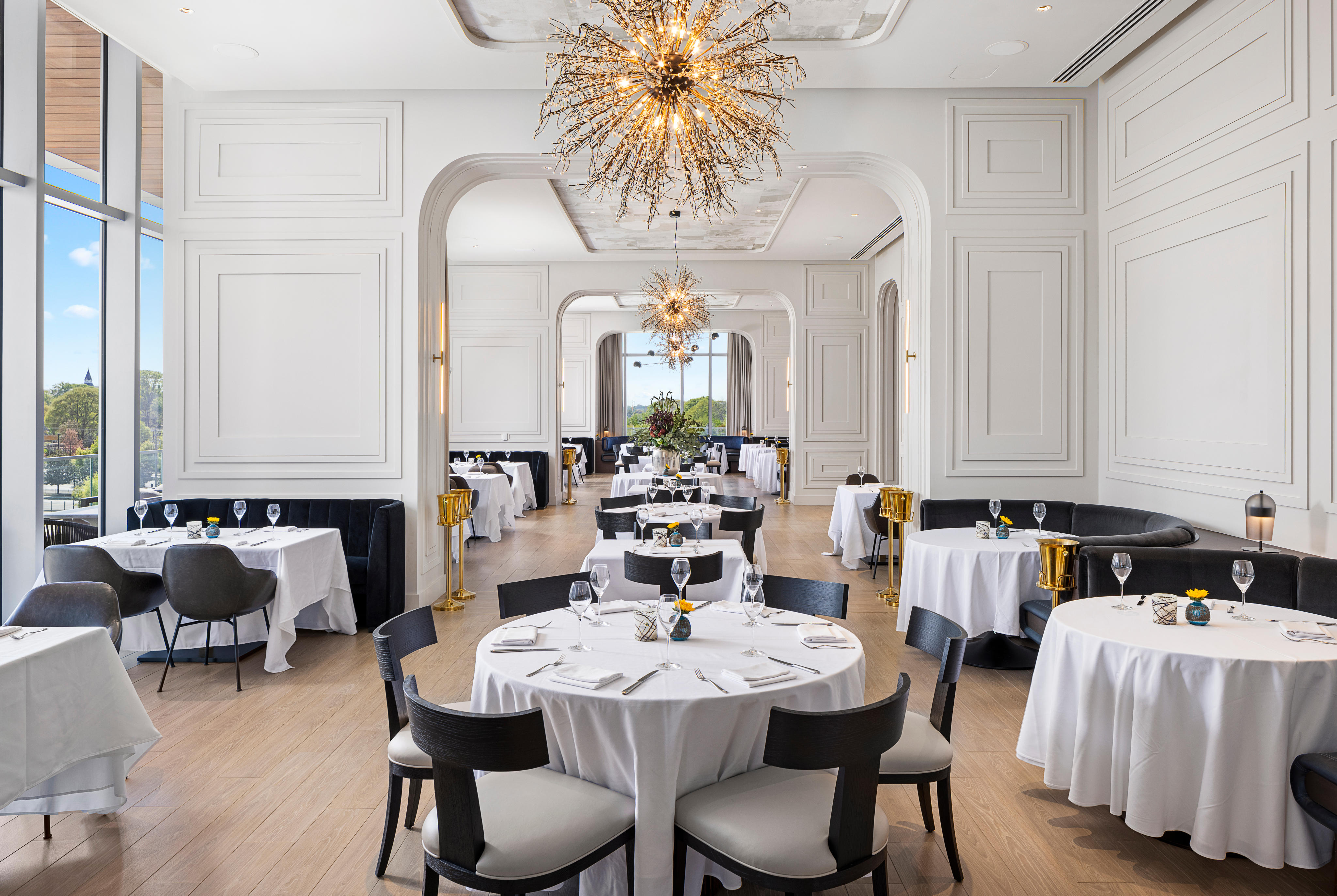 Elegant Italian dining room in Atlanta featuring round tables with white tablecloths, black chairs, and gold-accented chandeliers. Large windows provide natural light, enhancing the upscale ambiance.