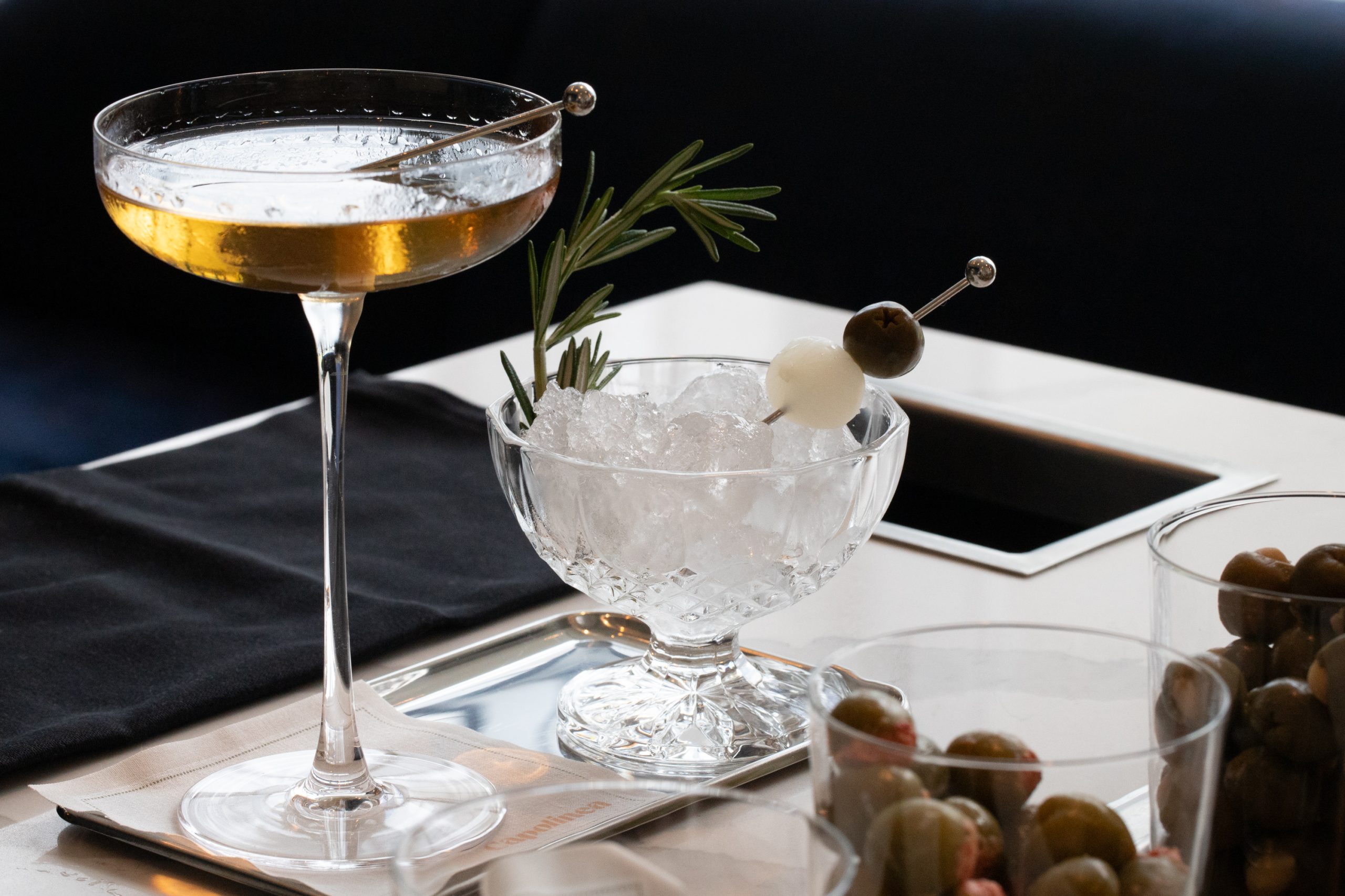 An upscale cocktail in a coupe glass with a rosemary garnish sits elegantly beside a dish of ice adorned with olives and skewered spheres on an Atlanta restaurant table.