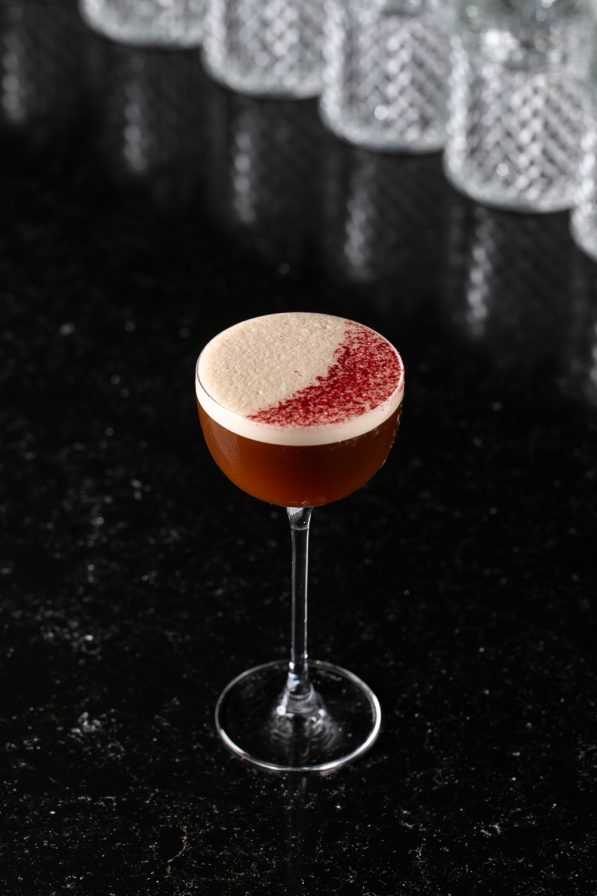 A cocktail in a coupe glass with a frothy top and a red garnish sits on a black surface, epitomizing upscale Italian dining. Crystal glasses blur into the background, enhancing the restaurant's sophisticated ambiance.