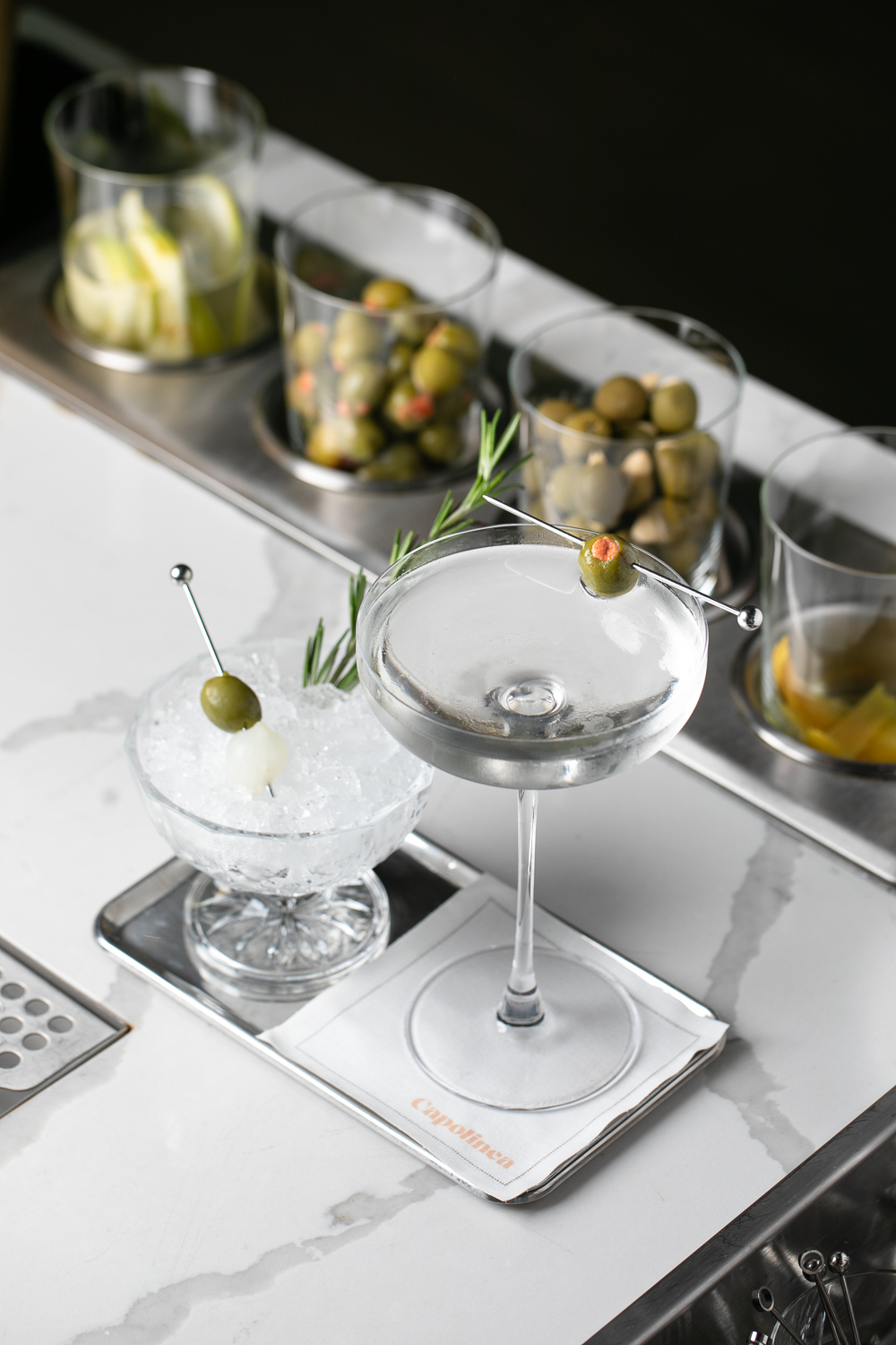 An upscale cocktail in an ice glass, garnished with olives and a rosemary sprig, sits elegantly on the bar counter of an Italian dining restaurant, with jars of garnishes adding to the sophisticated ambiance.