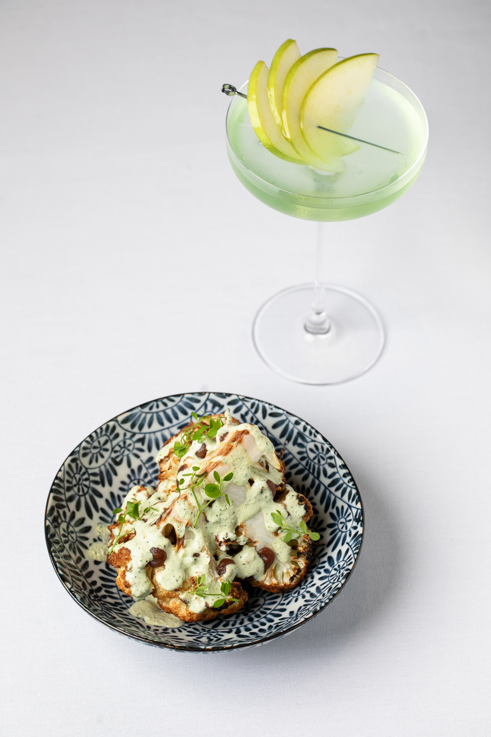 A plate of roasted cauliflower with creamy sauce and microgreens awaits in this upscale dining experience, perfectly paired with a green cocktail garnished with apple slices in a martini glass—an exquisite offering at an Italian restaurant in Atlanta.