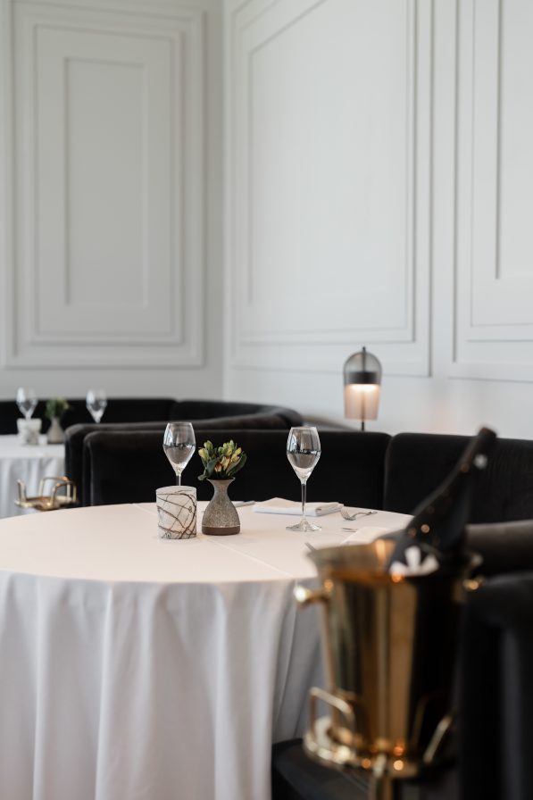 Elegant dining room with tables set for two, featuring white tablecloths, wine glasses, and a small potted plant. A wine cooler with a bottle is in the foreground—an exquisite taste of Italian dining right in the heart of Atlanta.