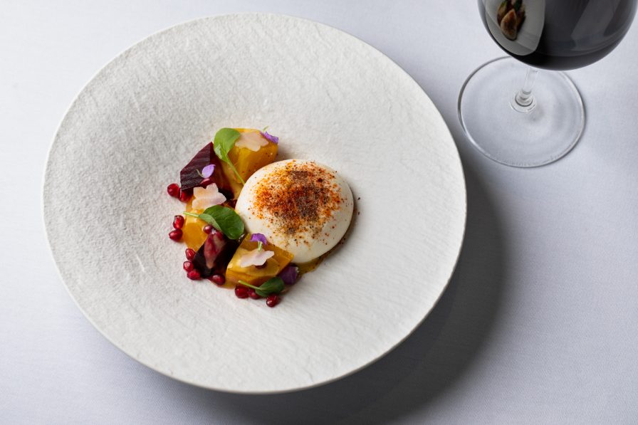 A white plate with a poached egg, garnished with spices, beetroot slices, greens, and pomegranate seeds embodies upscale elegance. An exquisite glass of red wine complements the dish, evoking the charm of Italian dining. Perfectly curated for a delightful Atlanta brunch experience.