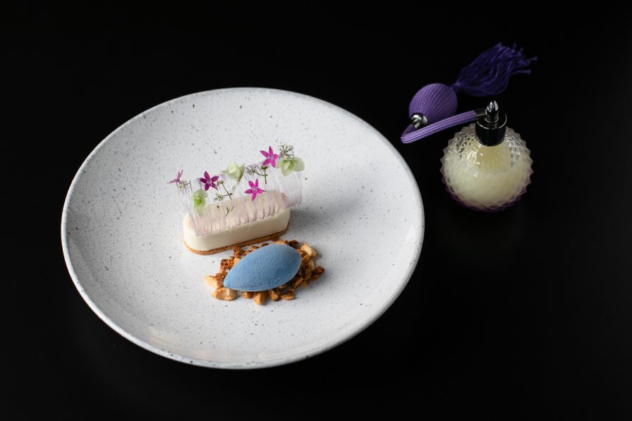 An upscale gourmet dessert featuring a delicate white mousse adorned with vibrant purple flowers and a blue scoop, elegantly presented on a white plate. Accompanied by a petite perfume bottle, this exquisite treat captures the essence of fine dining in an Atlanta restaurant setting.