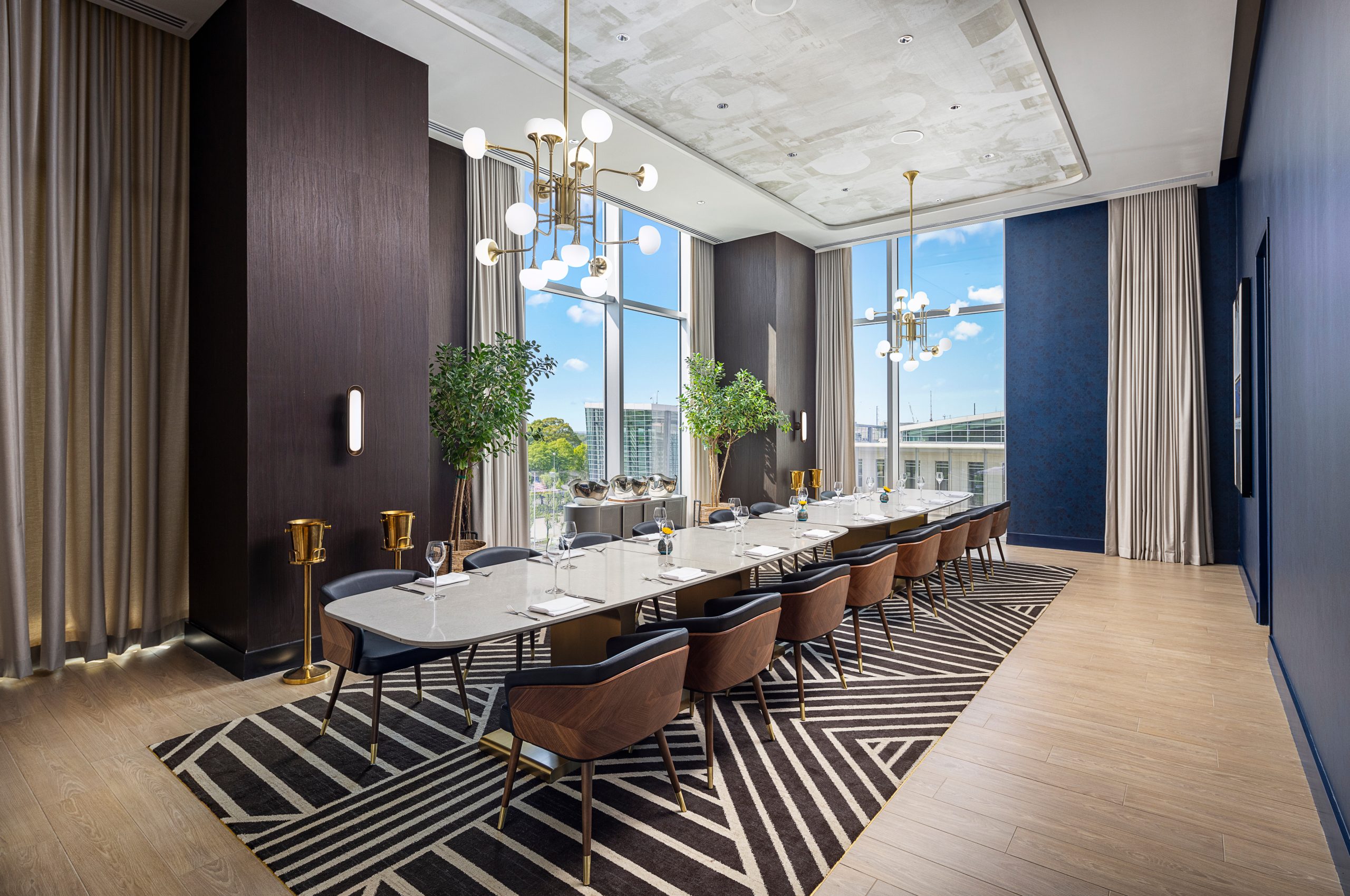 Upscale modern dining room in Atlanta featuring a long table with seating for twelve, a geometric rug, large windows, two decorative plants, and contemporary lighting fixtures.