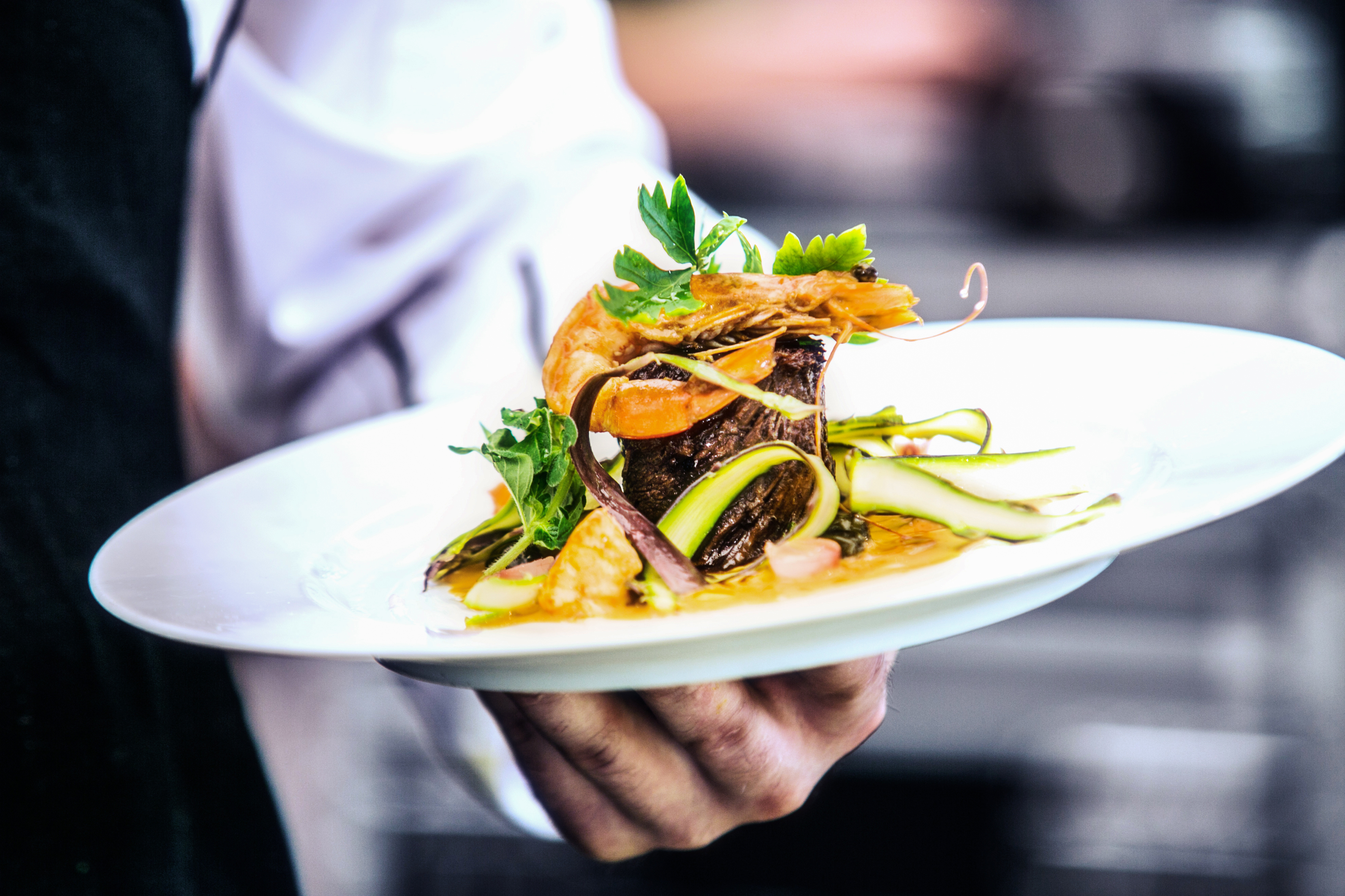 The chef presents a white plate with a gourmet dish of cooked shrimp, zucchini slices, and fresh herbs, embodying the essence of Italian dining. Discover this culinary delight at our restaurant in Atlanta.