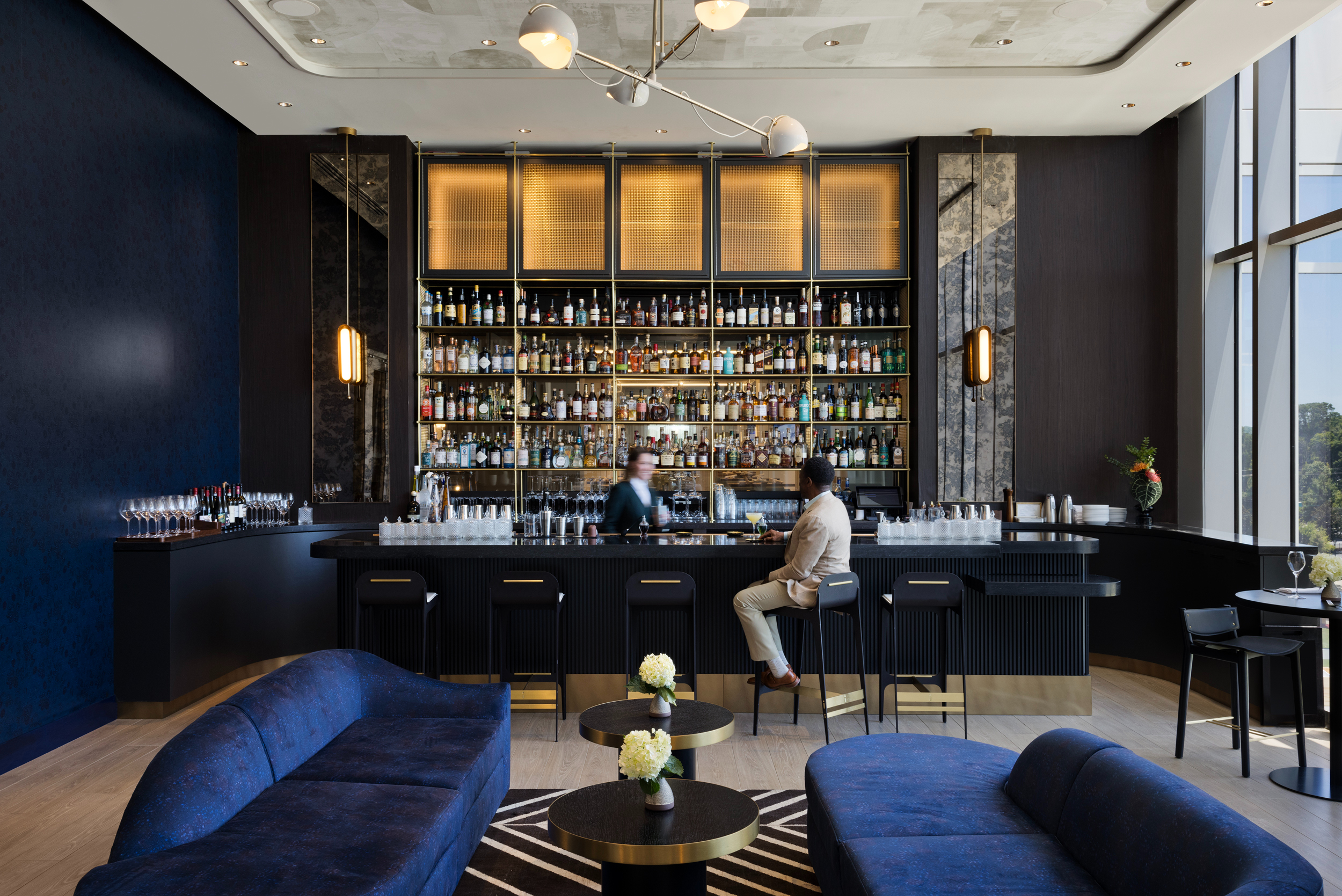 Two people sit at an upscale bar with a wall of bottles behind them. Located in Atlanta, the space features dark blue decor, a geometric rug, and large windows.