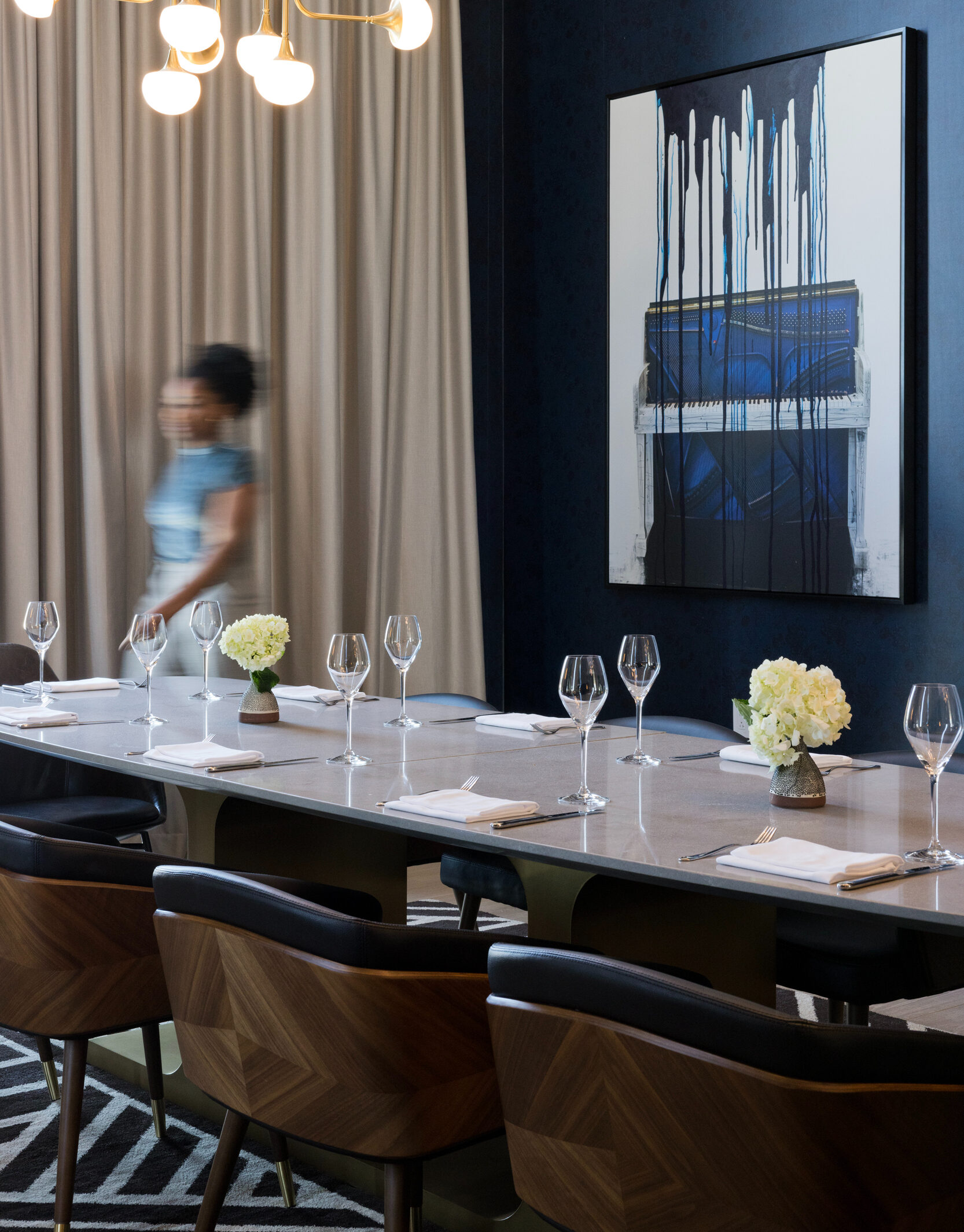 A modern dining room exudes the charm of Italian dining with an elegant table set with wine glasses and flowers. A blurred figure walks past, while a painting with dripping blue stripes adorns the dark wall, reminiscent of a chic restaurant in Atlanta.