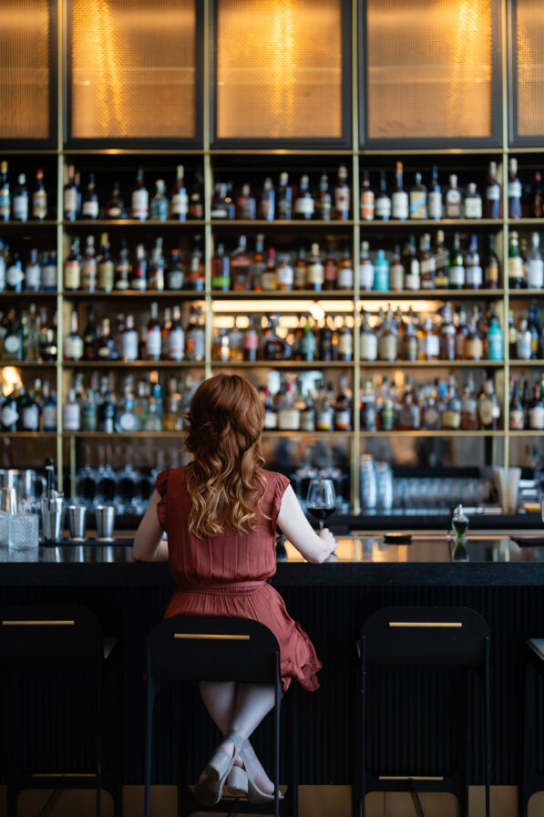 A woman with long red hair sits alone at a bar counter, facing shelves filled with bottles of alcohol.