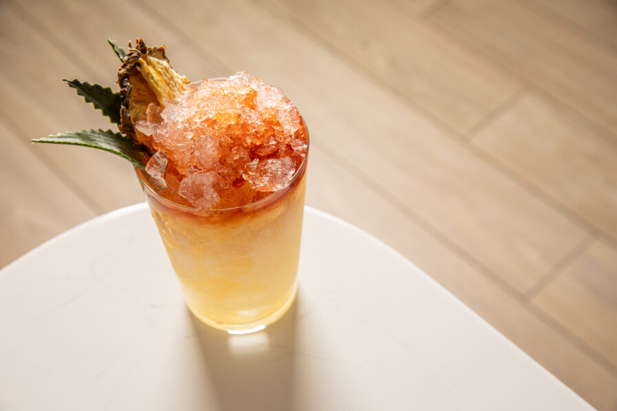 A glass of yellow cocktail with crushed ice, garnished with a pineapple slice and leaves, sits on a white surface with a wood floor background.