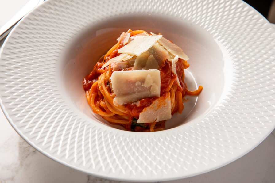 A bowl of spaghetti with tomato sauce, topped with several slices of parmesan cheese, served in a white textured dish.