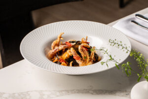 A white bowl contains a seafood pasta dish with mussels, fried soft-shell crab, and sliced red chili, garnished with herbs, set on a marble table.