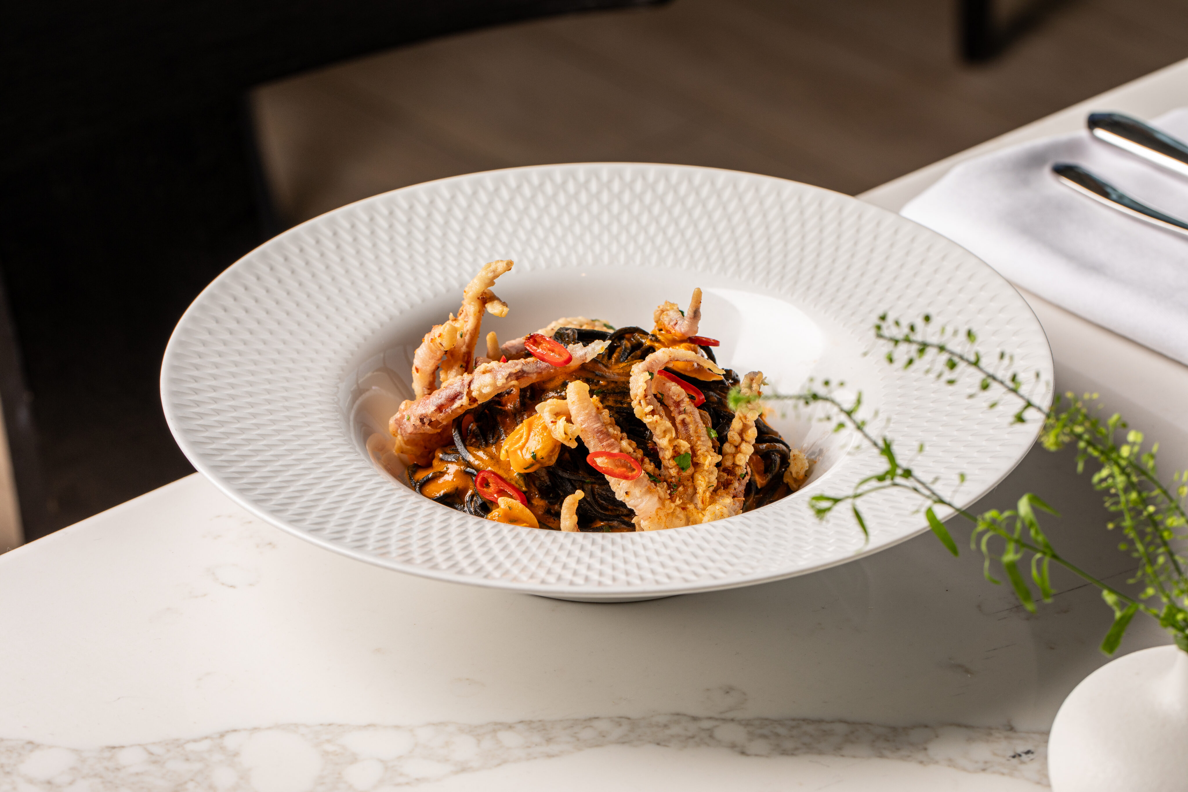 A white bowl contains a seafood pasta dish with mussels, fried soft-shell crab, and sliced red chili, garnished with herbs, set on a marble table.