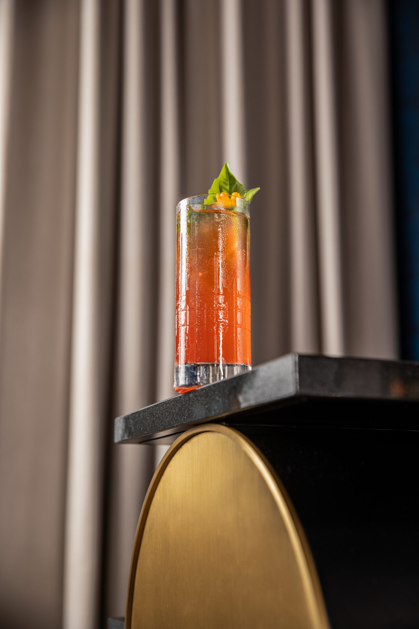 A tall glass of red-orange cocktail garnished with basil leaves sits on a black and gold table, with beige curtains in the background.