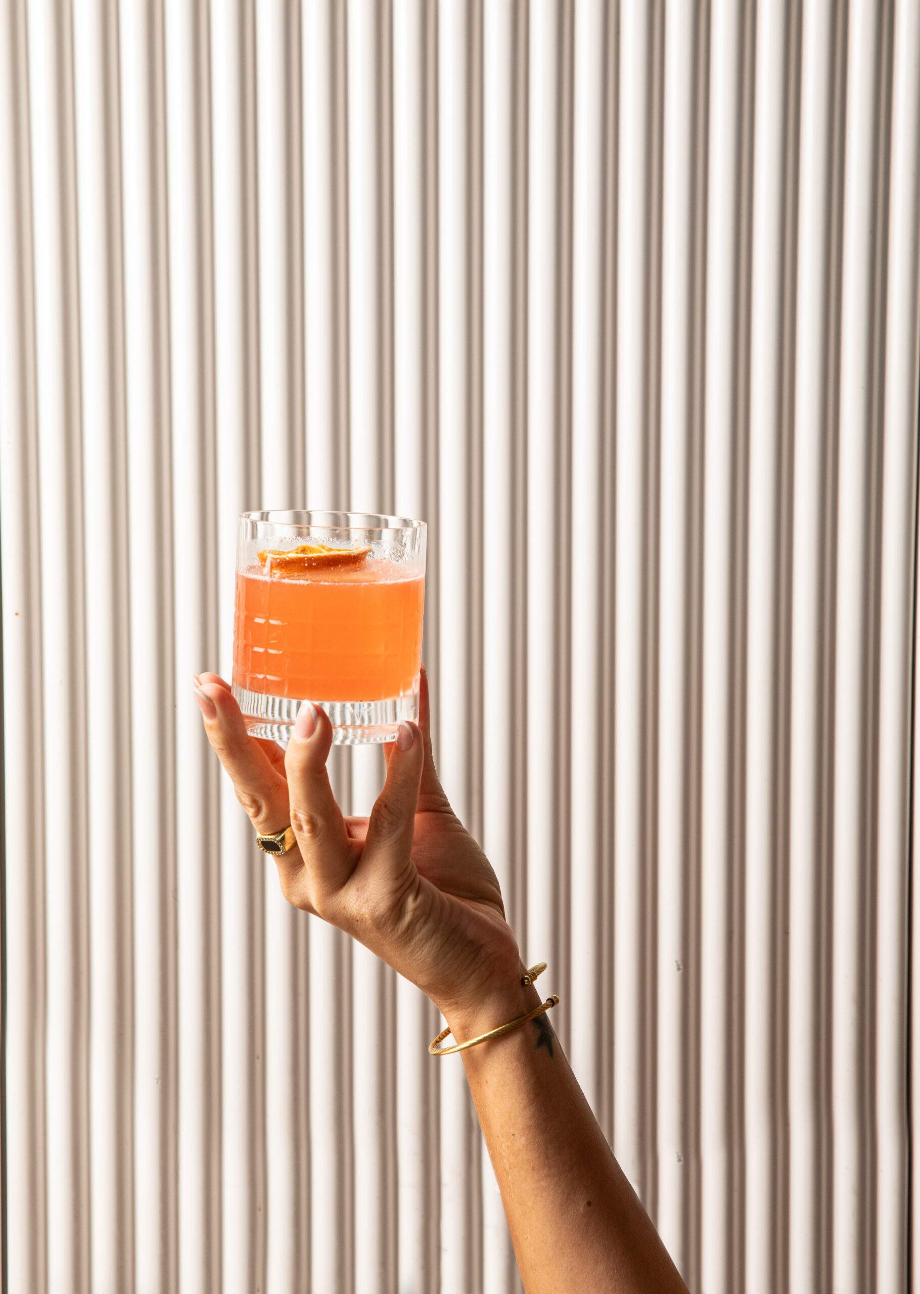 A hand with rings and a bracelet holds up a glass of orange-colored beverage in front of a vertically striped white background.