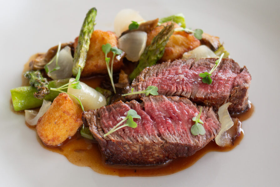 Sliced medium-rare steak with brown sauce, roasted potatoes, asparagus, onions, and microgreens on a white plate.