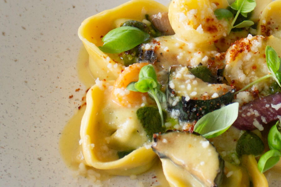 Close-up of tortellini pasta with creamy sauce, garnished with basil leaves, grated cheese, and slices of mushrooms on a white plate.